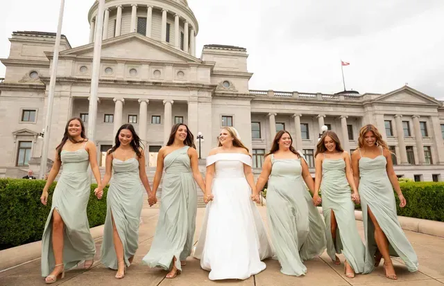 A bride and six bridesmaids in sage green dresses walk hand-in-hand outside a large, classical stone capitol building.