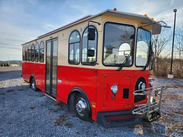 A red and cream-colored bus, designed to resemble a vintage trolley, parked on a gravel lot under a clear blue sky.