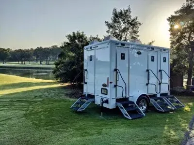 A white portable luxury restroom trailer with three entry doors and stairs parked on a grassy golf course near a pond.