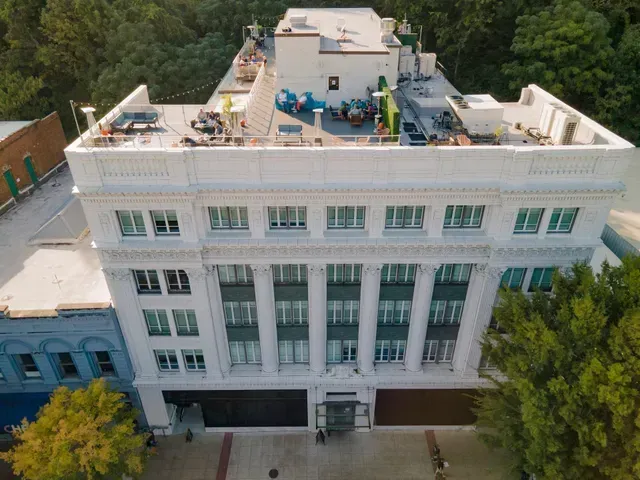 An aerial view of a white, multi-story historic building with columns and a populated rooftop terrace.