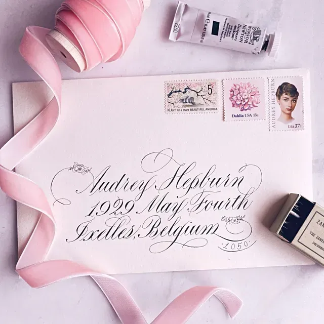 An envelope with elegant calligraphy addressed to Audrey Hepburn, decorated with vintage stamps, pink ribbon, and art tools.