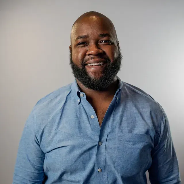 A smiling individual wearing a light blue button-down shirt against a light gray background.