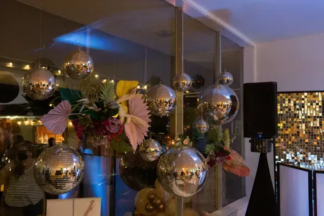 Hanging metallic silver disco balls and floral arrangements decorate an indoor event space with a DJ speaker in the back.