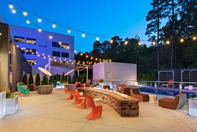 An outdoor lounge area at dusk, featuring a long stone fire pit, orange chairs, string lights, and a swimming pool.