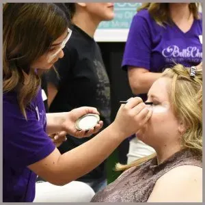 A makeup artist in a purple shirt applies eyeshadow to a client, holding a small palette in her other hand.