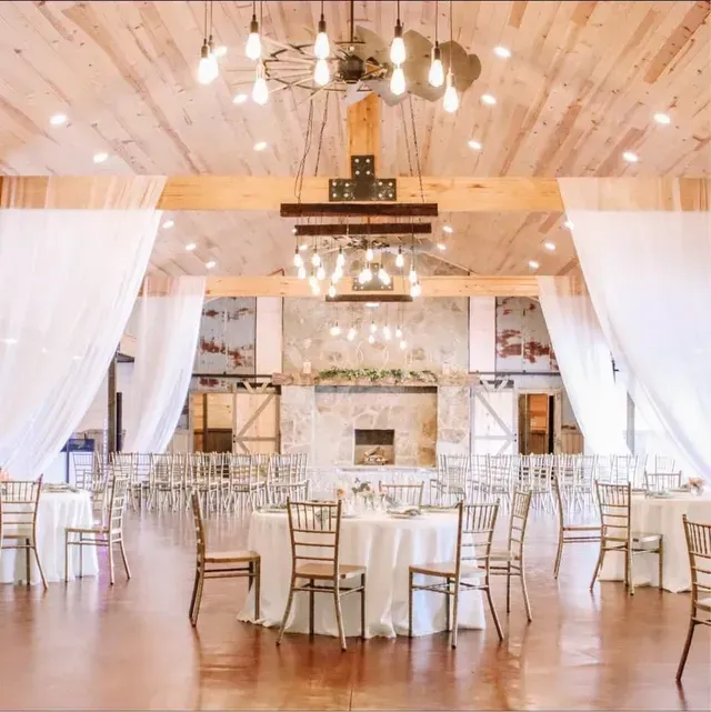 Rustic wedding venue with round tables, wooden beams, stone fireplace, and white drapery under a tiered wagon-wheel light.
