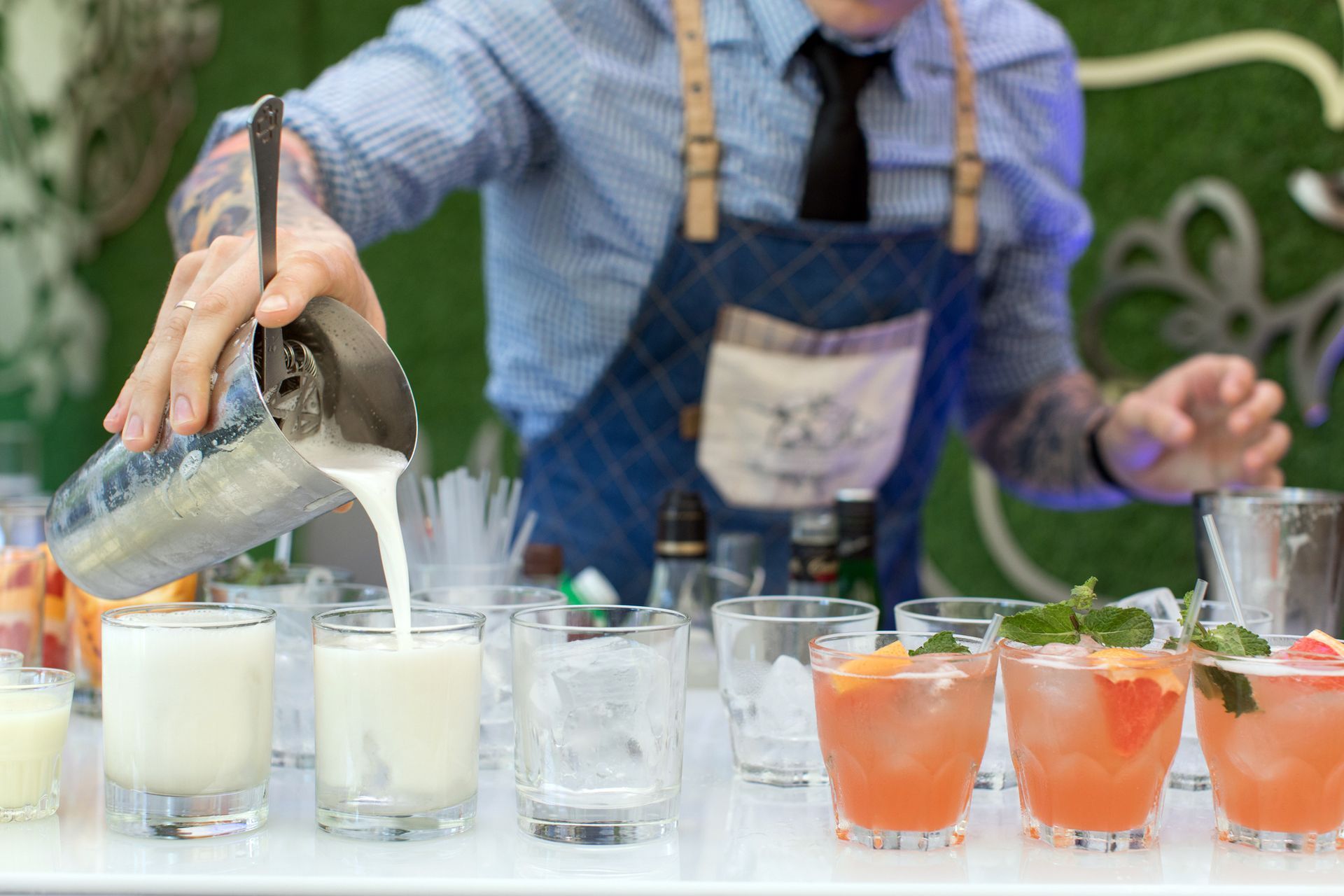 A bartender in a blue shirt and apron pours a white cocktail into a glass next to three pink cocktails with garnishes.