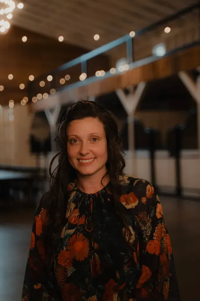 A person smiling at the camera, wearing a dark floral blouse in a dimly lit hall decorated with string lights.