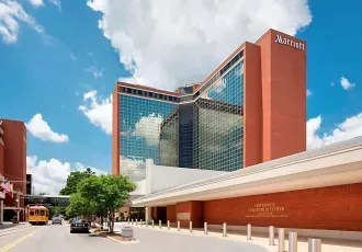 A tall red-brick Marriott hotel building with large glass windows under a bright blue sky, with a street and bus in front.