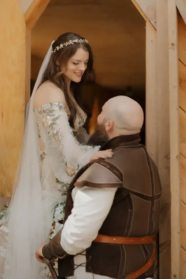 A person in an embroidered wedding gown and veil touches the shoulder of a person wearing medieval-style leather armor.