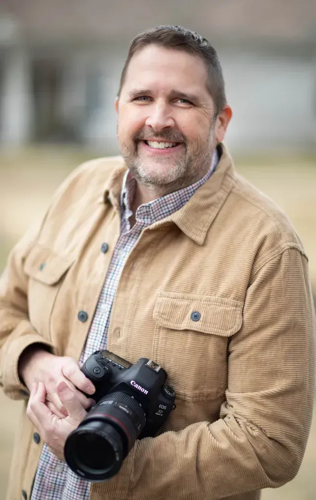A smiling person in a tan jacket holds a professional camera in front of a blurred outdoor background.
