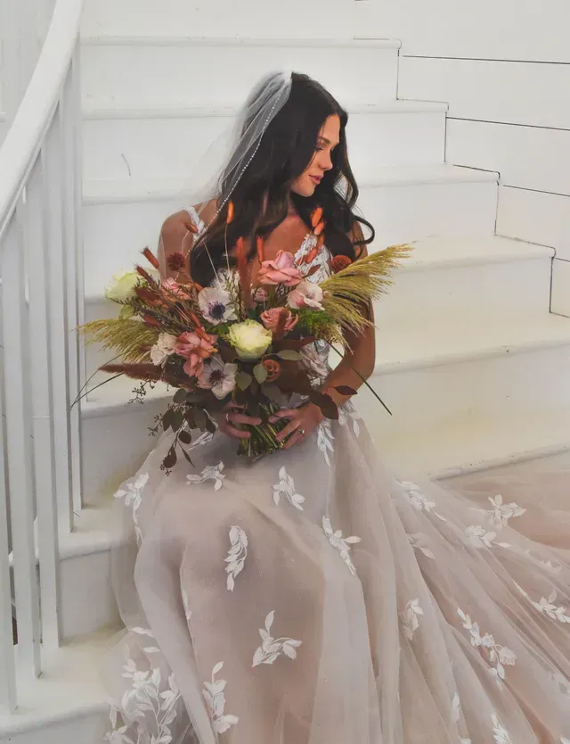 A person in a wedding dress and veil sits on white stairs, holding a large, colorful bouquet of flowers and dried grass.