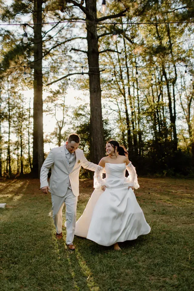 A bride and groom in wedding attire walk and laugh together on a grassy lawn beneath trees strung with outdoor lights.