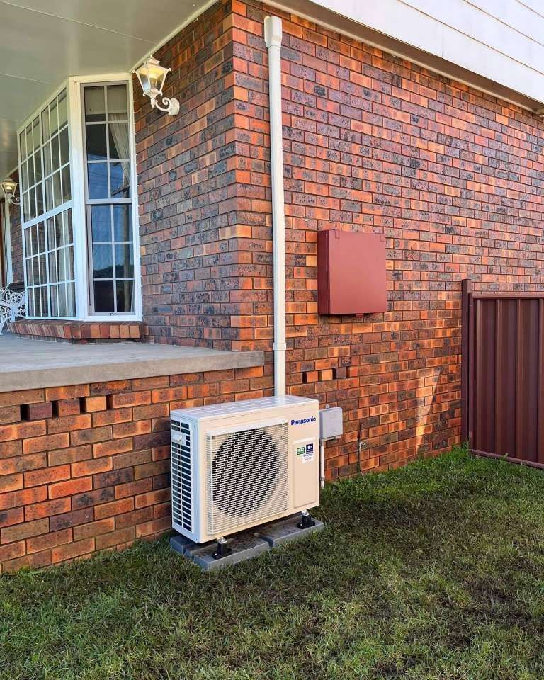 A White Air Conditioner Is Sitting On The Side Of A Brick House — Rapidcool Air Conditioning & Electrical In Dapto, NSW