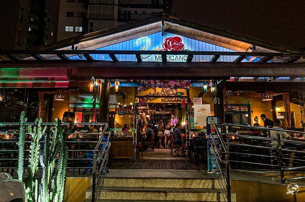 Entrada de restaurante à noite, bem iluminada, com pessoas jantando no interior. Decorações coloridas, teto azul, placa acima da entrada.