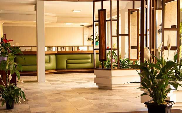 Área de recepção com assentos verdes, divisórias de madeira marrom, plantas e piso de azulejo.