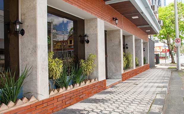 Fachada do edifício em tijolo e pedra, com grandes janelas, plantas e calçada.