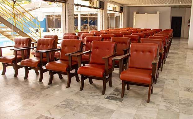 Fileiras de cadeiras vermelhas vazias em uma sala de conferências com piso de mármore e escadas ao fundo.