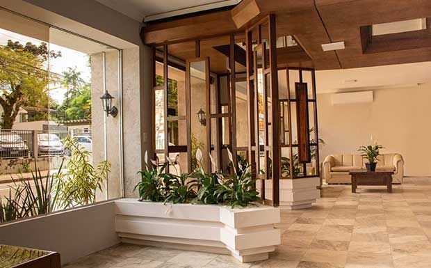 Hall de entrada com uma grande janela, divisória de madeira, plantas em um vaso, sofá bege e piso de mármore.