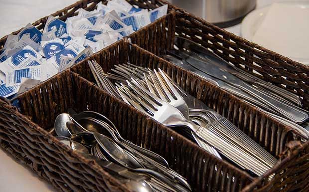 Organizador de talheres com colheres, garfos, facas e sachês de açúcar em uma cesta de vime.