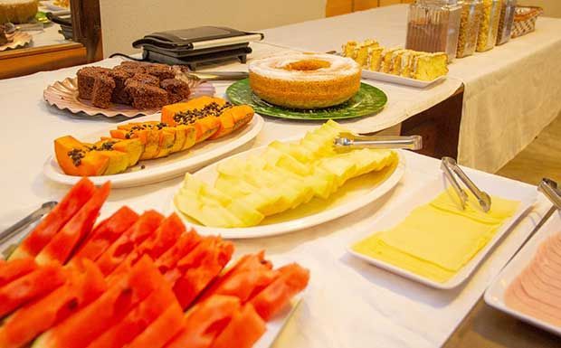 Buffet de café da manhã com melancia, mamão, bolo e queijo sobre uma toalha de mesa branca.