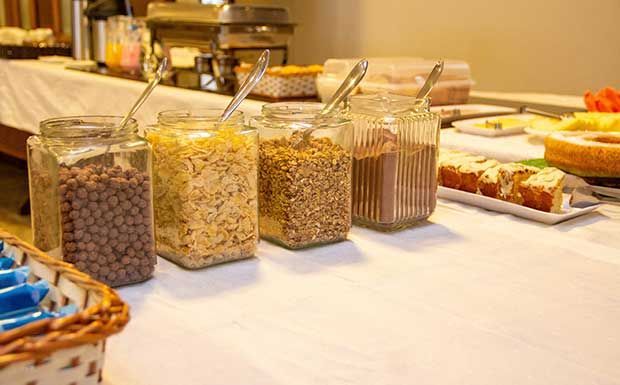 Buffet de café da manhã: Potes de cereal, com colheres, sobre uma toalha de mesa branca. Outros itens alimentícios visíveis.