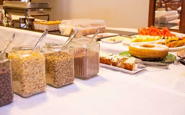 Buffet de café da manhã com potes de cereais, pão, bolo e frutas sobre uma toalha de mesa branca.