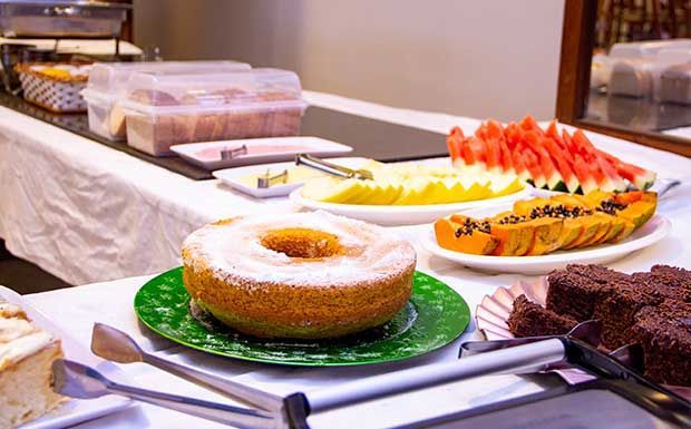 Buffet de café da manhã com bolo, fatias de frutas e doces sobre toalha de mesa branca.