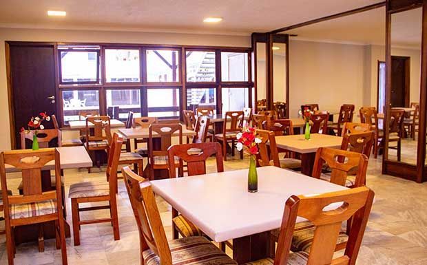 Interior de restaurante com mesas e cadeiras de madeira, paredes brancas e grandes janelas.
