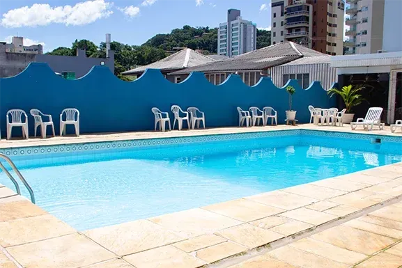 Piscina azul rodeada por cadeiras brancas, com uma parede azul e edifícios ao fundo num dia ensolarado.
