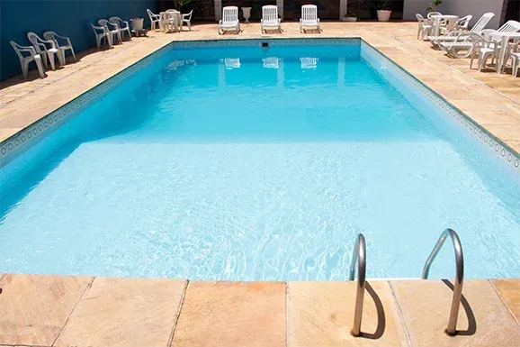 Piscina retangular com água azul cristalina e escada de aço. Espreguiçadeiras e cadeiras rodeiam a piscina.