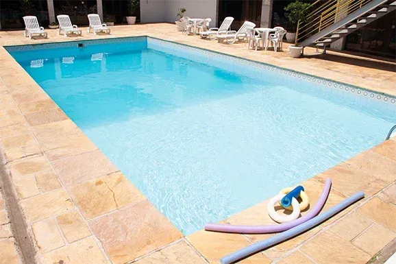 Piscina com água azul, rodeada por ladrilhos de pedra. Boias repousam na borda.