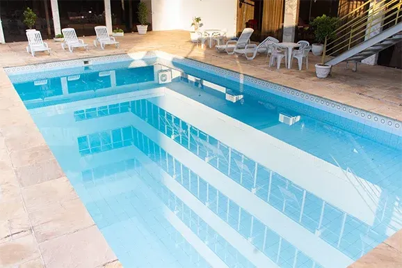 Piscina com água cristalina que reflete um edifício; espreguiçadeiras e mesas no deck.