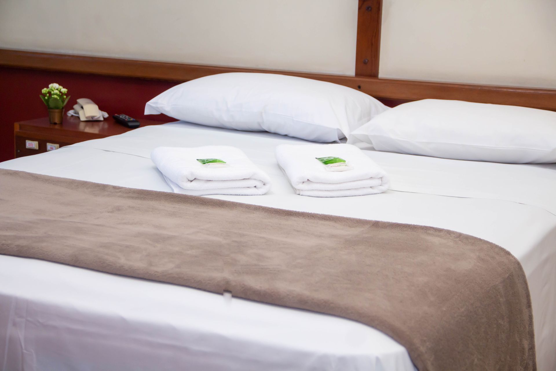 Quarto com lençóis brancos, manta marrom, duas toalhas e uma mesa de cabeceira.