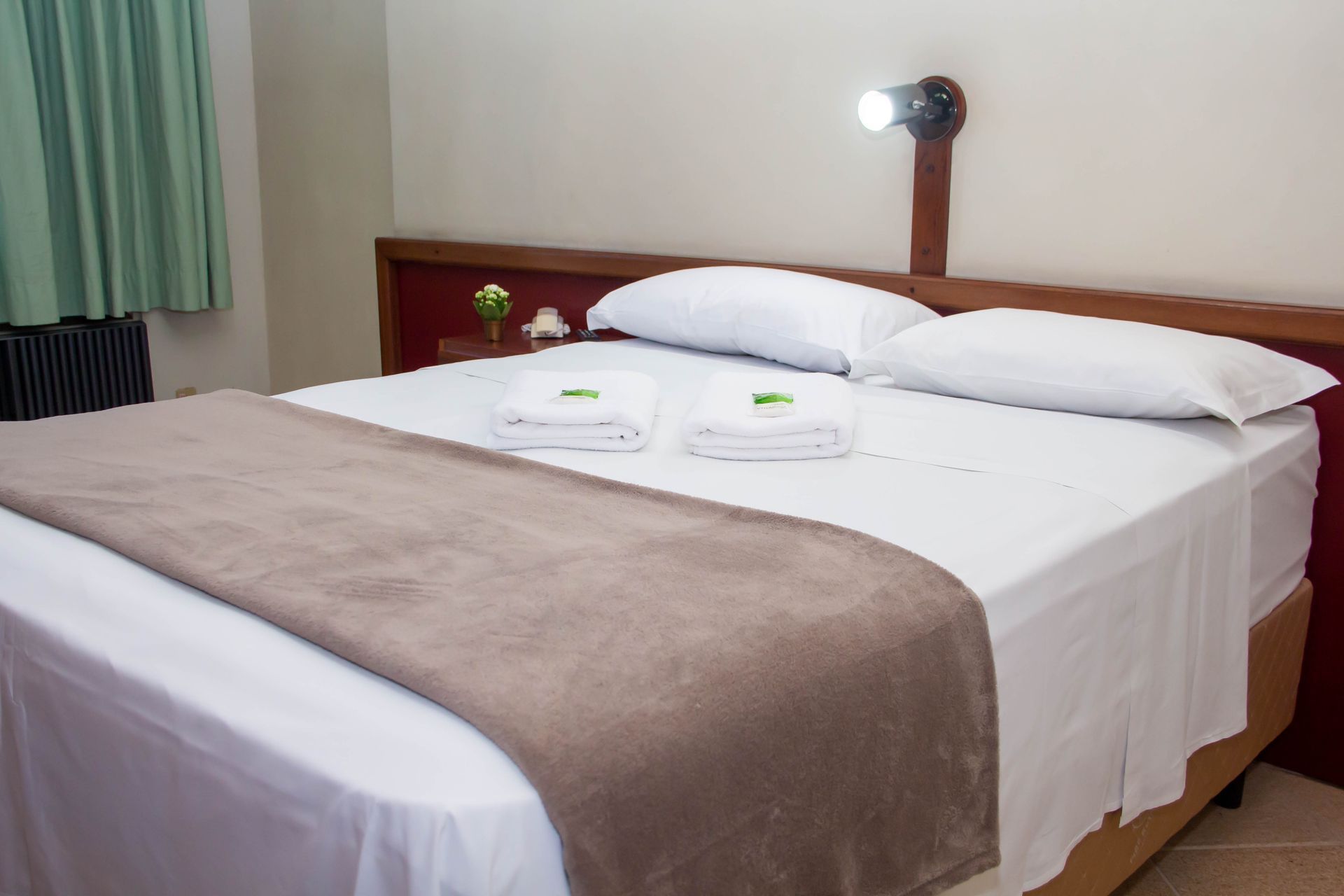 Quarto de hotel com cama, lençóis brancos e cobertor marrom. Duas toalhas enroladas e uma luminária de parede.