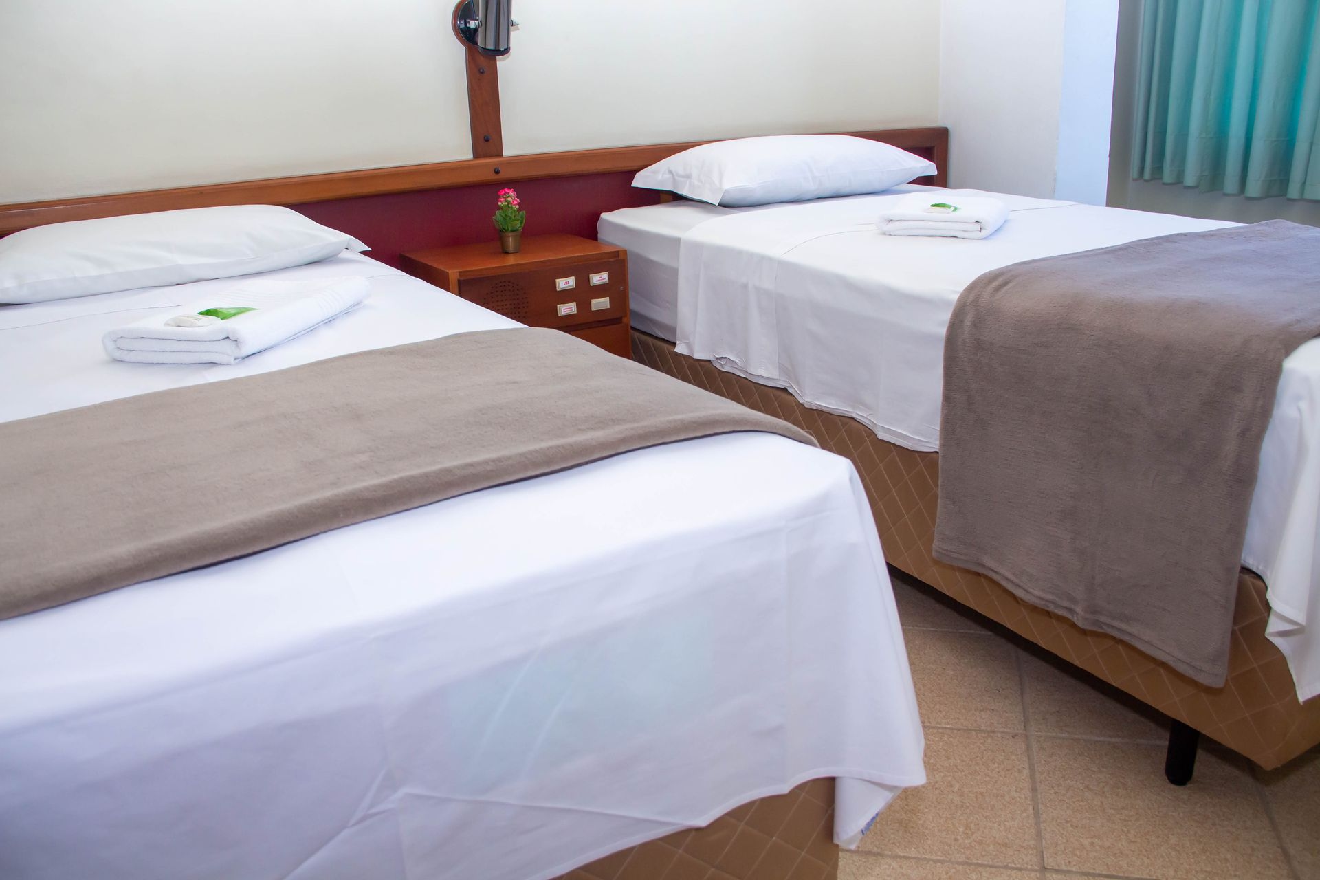 Duas camas de solteiro em um quarto com lençóis brancos, cobertores marrons e uma pequena mesa de cabeceira.