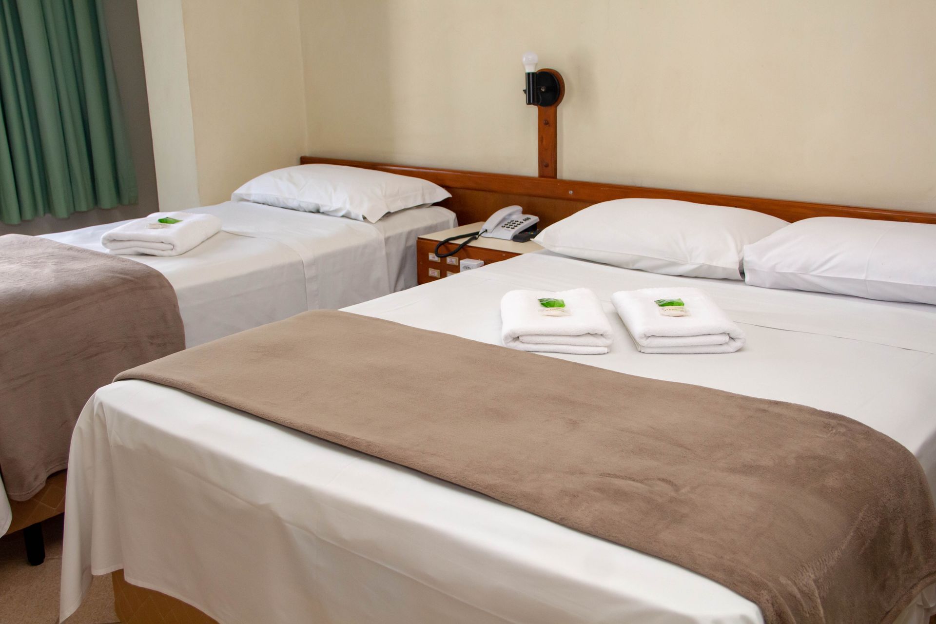 Quarto de hotel com uma cama de casal e uma cama de solteiro; lençóis brancos, manta marrom e toalhas dobradas.