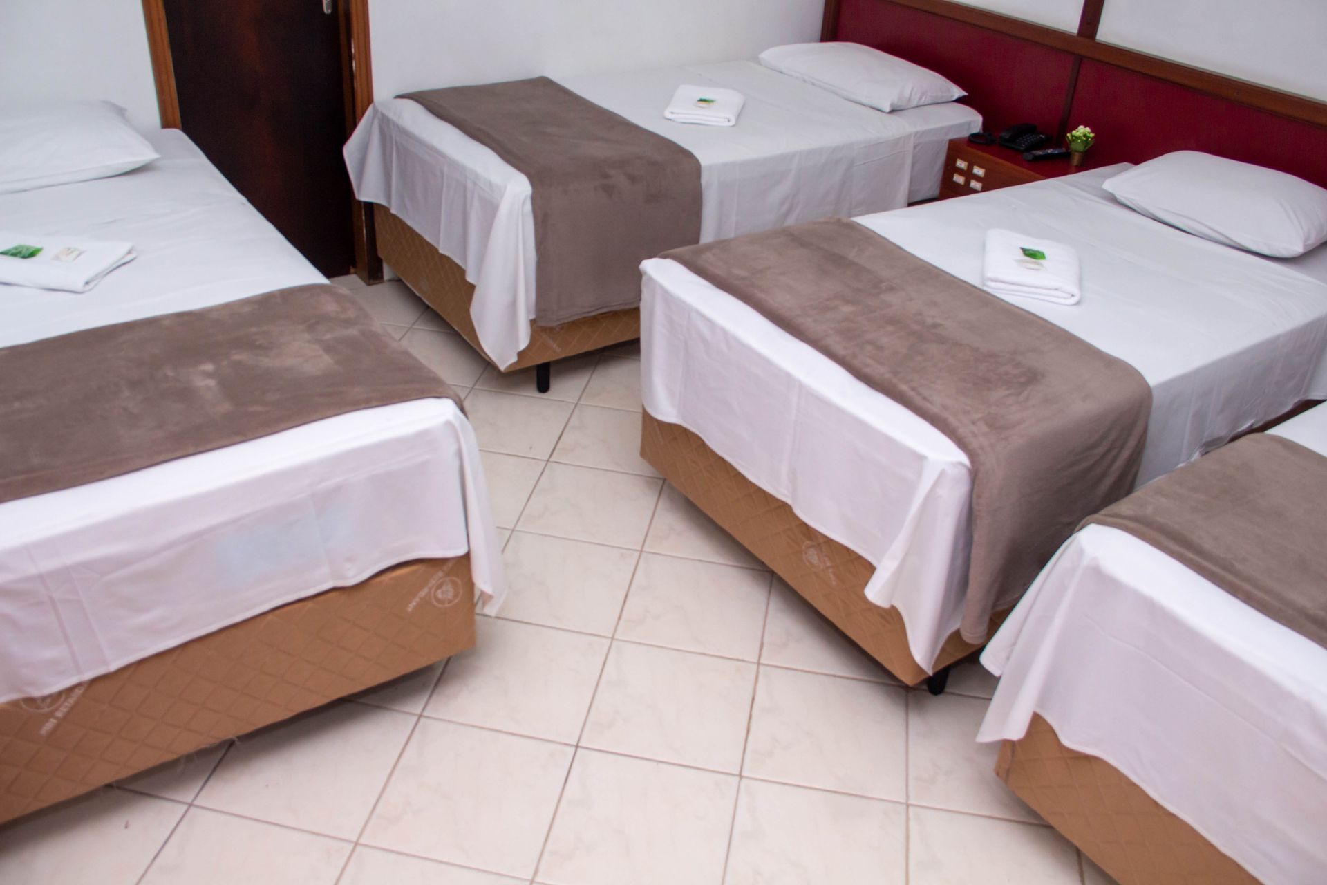 Quatro camas de solteiro em um quarto de hotel, cada uma com um lençol branco, cobertor marrom e travesseiro, sobre um piso de azulejos.