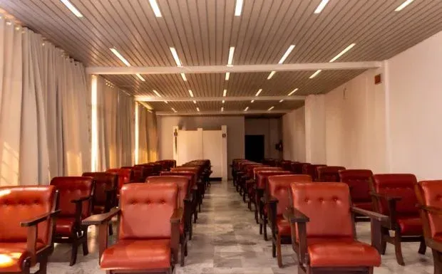 Fileiras de cadeiras vermelhas estão dispostas de frente para uma porta em uma sala com teto quadriculado, cortinas e paredes brancas.