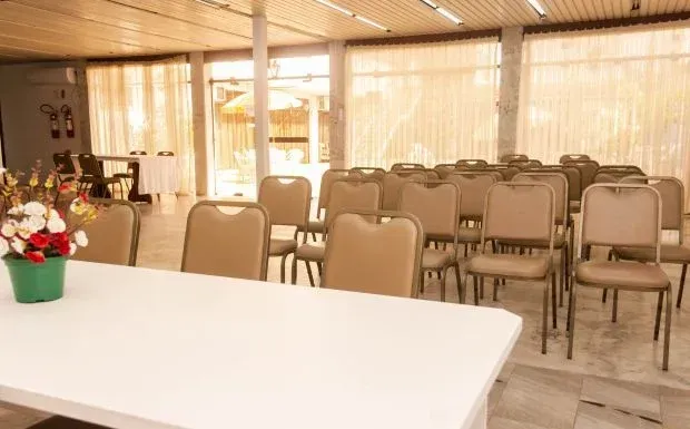 Sala de reuniões com fileiras de cadeiras bege, uma mesa branca e um arranjo floral.