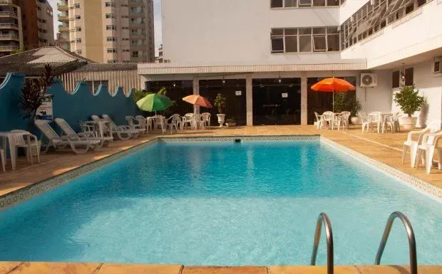 Área da piscina com água turquesa, espreguiçadeiras, mesas com guarda-sóis e um prédio ao fundo.