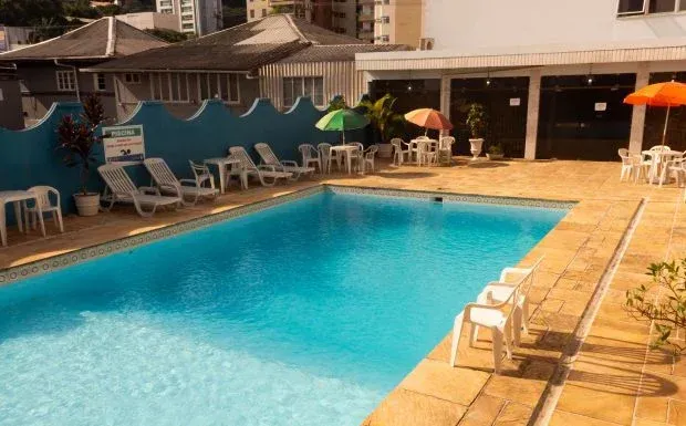 Piscina com espreguiçadeiras e mesas. Prédios ao fundo. Água azul brilhante sob a luz do sol.