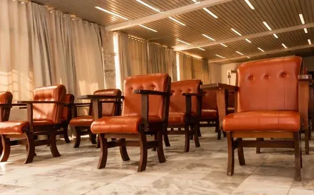 Fileiras de poltronas de couro marrom em uma sala com cortina ao fundo e luminárias.