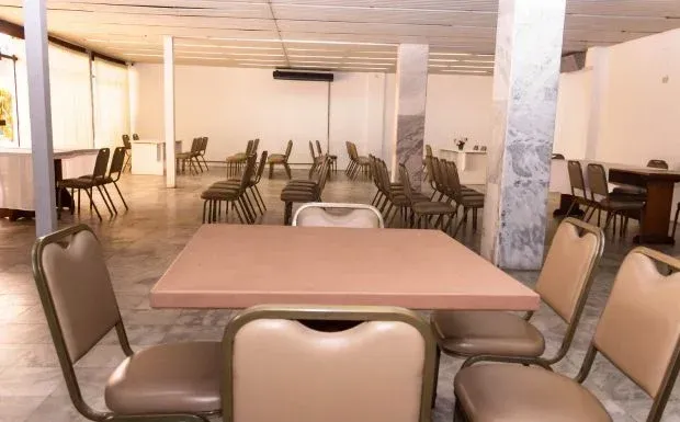 Sala vazia e com poucos móveis, composta por mesas e cadeiras. Tons de bege e cinza.