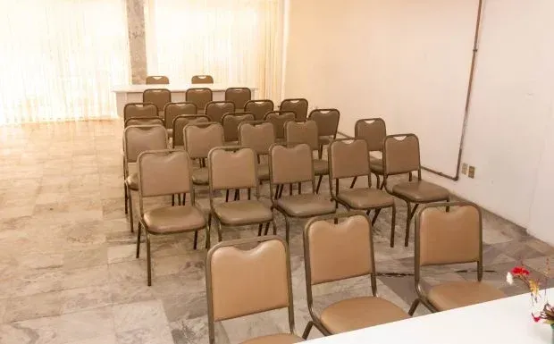 Sala de conferências vazia com fileiras de cadeiras bege dispostas em frente a uma mesa. Paredes brancas, iluminação forte.