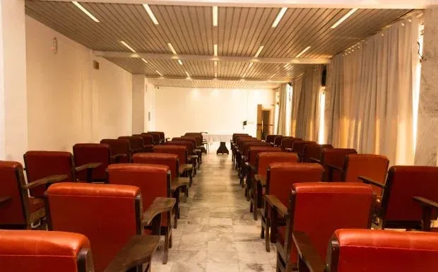 Auditório vazio com fileiras de cadeiras vermelhas voltadas para uma tela em branco; luzes brilhantes no teto.