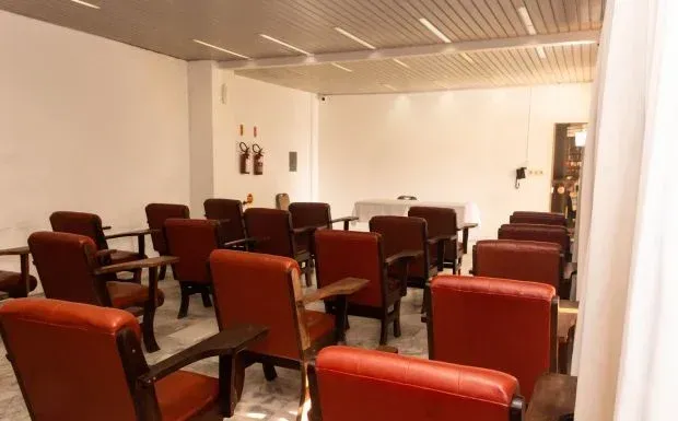 Auditório vazio com fileiras de cadeiras vermelhas, paredes brancas e uma pequena mesa.