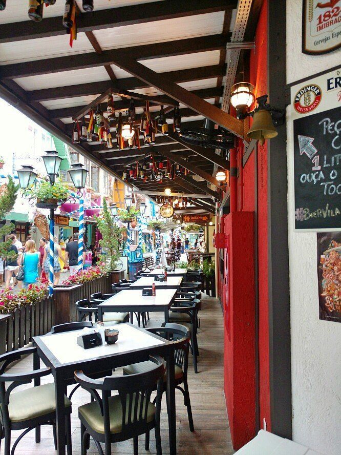 Restaurante ao ar livre com mesas, cadeiras e decoração suspensa. Parede vermelha, área externa com vista para a rua.