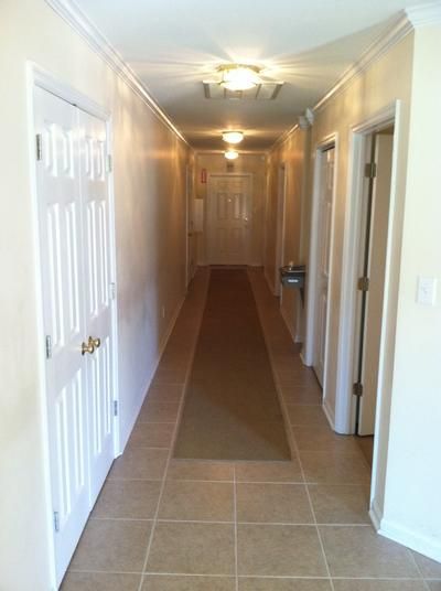Long hallway with off-white walls, tiled floor, carpet runner, doors on each side, and recessed lighting.