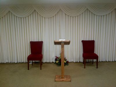 Funeral home interior: two red chairs, wooden podium, flowers, and draped white curtains.